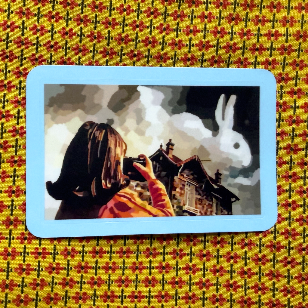 A square art print with rounded corners featuring an illustration of a little girl taking a photo of a cloud shaped like a rabbit in a dark sky above houses, set against a yellow and red patterned fabric background.